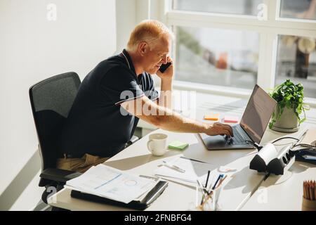 businessman sitting at office desk and talking on mobile phone. Male business professional using laptop and making phone call. Stock Photo