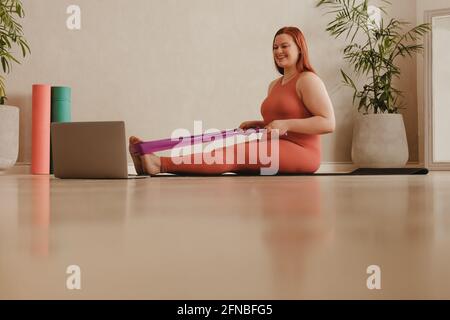 Woman doing stretching exercise using resistance band and looking at laptop. Smiling fit woman watching video on laptop and doing stretching workout. Stock Photo
