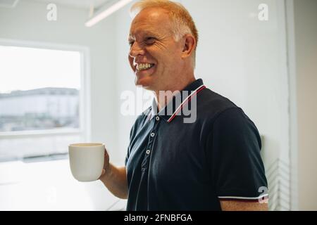 Senior businessman smiling while taking a coffee break. Happy mature man with coffee mug standing in office looking away. Stock Photo