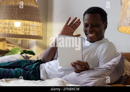 Black man with tablet resting in bed greeting waving hand. Stock Photo