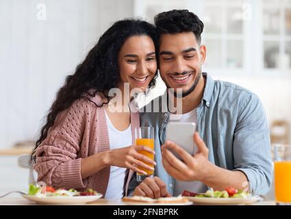 Portrait Of Happy Arab Couple Watching Videos On Smartphone And Having Breakfast Stock Photo