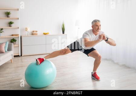Athletic mature man doing side lunges, using fitness ball, working out at home in living room interior Stock Photo