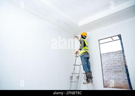 painter man painting the wall in an unfinished building, with paint brush and bucket, isolated on big empty space with ladder at the construction site Stock Photo