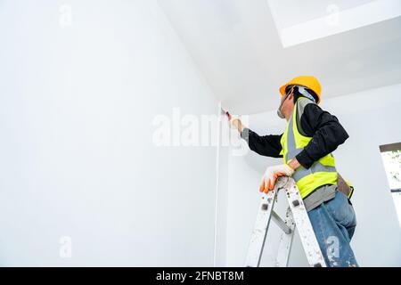 painter man painting the wall in an unfinished building, with paint brush, isolated on big empty space with wooden ladder at the construction site Stock Photo
