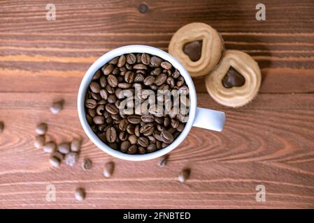 Coffee grains or beans in a ceramic mug with biscuits on wooden table. Top view of roasted  coffee seeds. Stock Photo