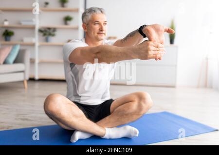 Sporty senior man doing stretching for hand exercises, sitting on mat in living room interior, free space Stock Photo
