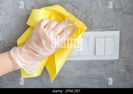 female hand clean light switch with cloth on gray concrete wall. Woman in glove disinfecting surfaces by yellow rag. The new normal Covid coronavirus Stock Photo