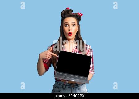 Shocked pinup woman in retro outfit pointing at laptop with empty screen on blue background Stock Photo