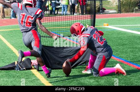 Rear view of a high school football field goal kicker on the sidelines during a game warming up kicking into a net with his holder. Stock Photo