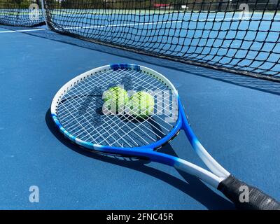 A Tennis racket on the ground with two ball under the racket on a blue court in front of the net Stock Photo