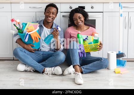 Smiling black couple sitting on floor, holding cleaning tools Stock Photo