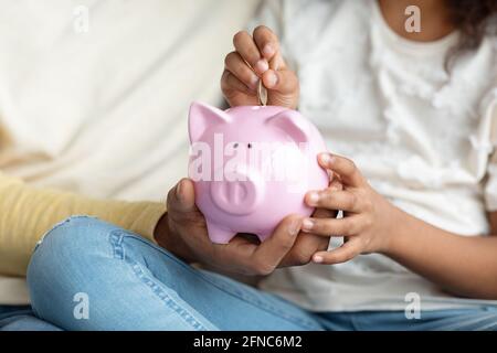 Black girl and father putting coin into piggy bank, cropped Stock Photo