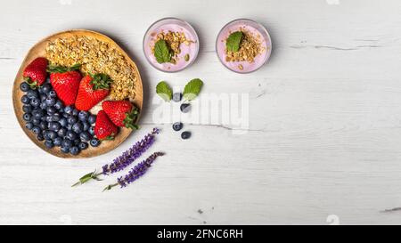 Top view of two bowls of berries yogurt with granola and oats and a wood plate of strawberries and blueberries for healthy breakfast on a rustic white Stock Photo