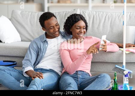 Black lovers with cleaning tools sitting on floor, using smartphone Stock Photo