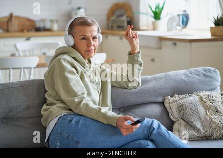 Movie time. Portrait of beautiful middle aged woman in headphones with remote control in hand watching tv while sitting and relaxing on sofa, spending leisure time at home. Selective focus on female Stock Photo