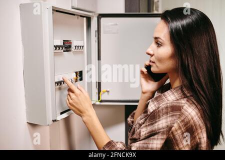 Young woman with mobile phone by ear standing by open electrical panel and calling repair service Stock Photo