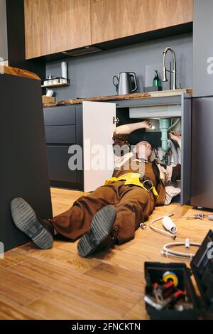 Young plumber lying on the floor while repairing kitchen sink Stock Photo