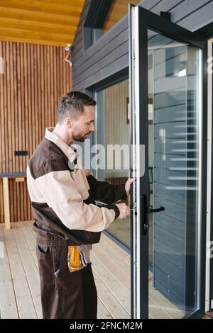Young repairman standing in front of a glass door while repairing lock Stock Photo