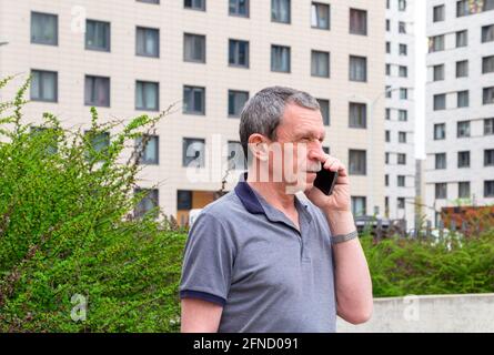 Adult caucasian senior man in a T-shirt talking on a smartphone in the city in spring or summer against the background of buildings. Selective focus. Stock Photo