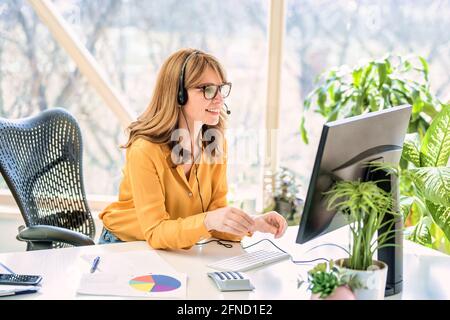 Mature businesswoman in headset speaking by conference call while looking at computer. Home office. Stock Photo