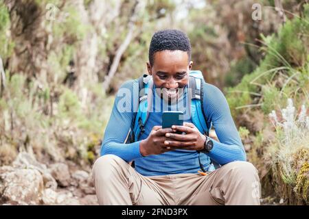 Young African ethnicity man video chatting via cellphone with somebody and cheerfully laughing as he having a hiking walk in the forest. Happy people, Stock Photo