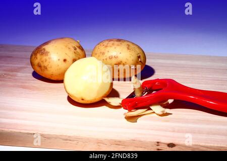Raw potatoes and a peeler on a wooden chopping board Stock Photo