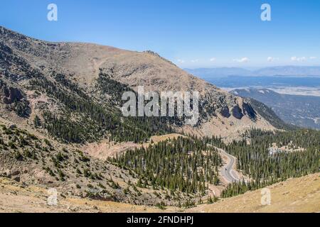 View from above of winding mountaind road near tree line with cars and a parking lot and a far view of valley and lake Stock Photo