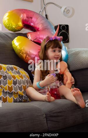 A Cute Baby Girl Sitting On A Sofa With A Colourful Balloon And A Doll Stock Photo