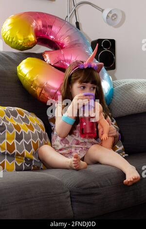 A Cute Baby Girl Sitting On A Sofa With A Colourful Balloon And A Doll Stock Photo