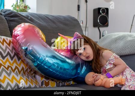 A Cute Baby Girl Sitting On A Sofa With A Colourful Balloon And A Doll Stock Photo