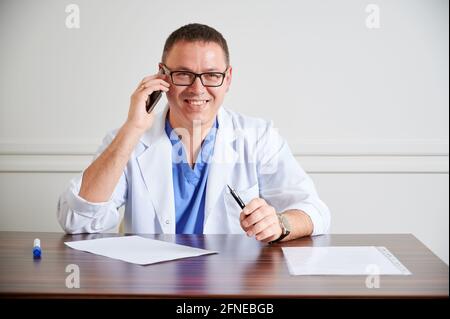Joyful man surgeon in glasses smiling while sitting at the table with papers. Male physician holding pen and talking on cellphone in clinic. Concept of medicine, communication and healthcare workers. Stock Photo