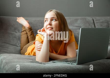 Pensive Caucasian beautiful woman lies on sofa with laptop. Portrait of dreamy teenager girl thinking behind laptop at home in living room with Stock Photo