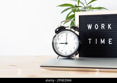 Classic vintage black alarm clock, laptop, potted plant and board sign 'work time' on wooden desk with white wall background and copy space for text. Stock Photo