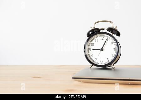 Classic vintage black alarm clock and laptop on wooden table with white wall background and copy space for text. Time and work concept. Stock Photo