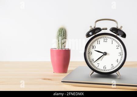 Classic vintage black alarm clock, laptop and small potted cactus on wooden table with white wall background and copy space for text. Time and work co Stock Photo