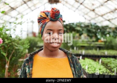 Portrait of African young woman with colorful headdress on her head smiling at camera while working in greenhouse Stock Photo