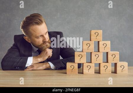 Businessman in suit looking at pyramid of cubes with question marks and thinking of answer Stock Photo