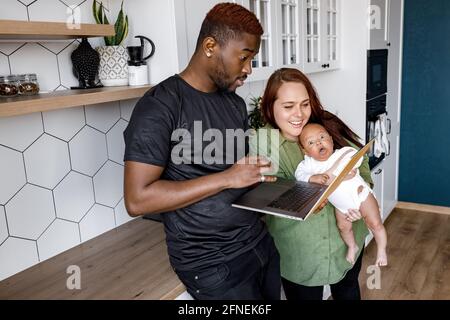 Smiling multiethnic parents relax at home with cute little biracial son using laptop together, happy international family with small boy child stay in Stock Photo