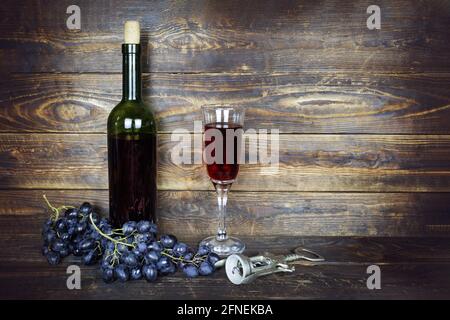 Dark green bottle and transparent wineglass with red wine and bunch of grapes on brown wooden plank surface, corkscrew nearby Stock Photo