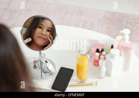 Pretty smiling young woman looking at magnifying mirror when applying undereye patches and lotions Stock Photo