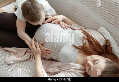 A little boy lies on the couch with his pregnant mother and hugs her tenderly. Stock Photo