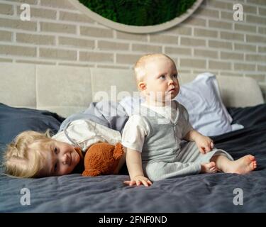 Cute baby boy with blue eyes sitting on bed at home and looking at something while playing and spending time with her older sister, happy little girl lying near brother and hugging teddy bear toy Stock Photo