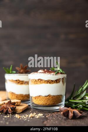 Layered dessert in glass jar with cookie crumble and whipped cream decorated with rosemary and anise, dark background. No bake cheesecake, trifle or p Stock Photo