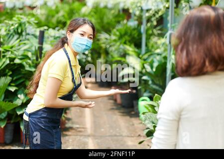 Pretty young woman in medical mask welcoming customers in plant nursery Stock Photo