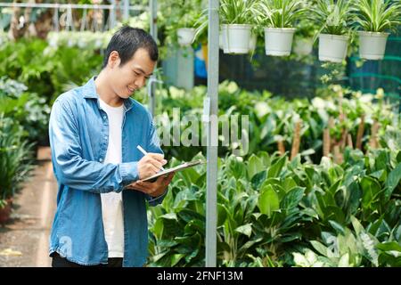 Smiling young man working at plant nursery, counting plants and taking notes in document Stock Photo