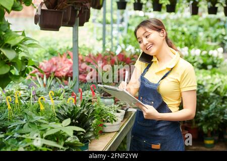 Smiling pretty gardening center worker talking on phone with customer and taking notes in document Stock Photo