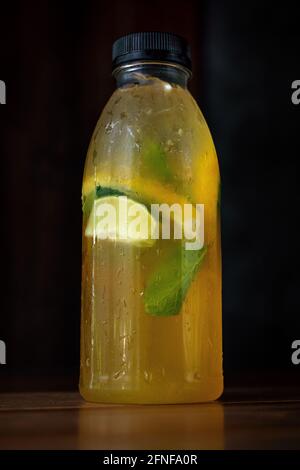 Cold lemonade with slices of lemon, orange and ginger with mint leaves in a closed bottle. Cold refreshing drink or beverage with ice on wooden table. Dark background. Vertical shot. Stock Photo
