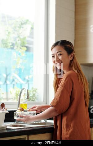 Pretty joyful young woman washing dishes in kitchen sink, turning to camera and smiling Stock Photo