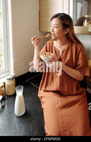 Lovely young woman sitting on kitchen counter, eating sweet cereals and looking on the street through window Stock Photo