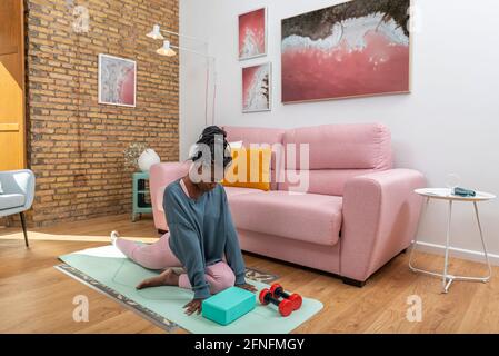 Black female in activewear stretching legs before workout at home while sitting on mat Stock Photo